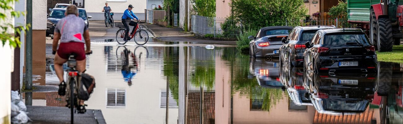Radler fahren durch eine überflutete Straße. Im Bereich der Gemeinde Baar-Ebenhausen war ein Damm an zwei Stellen geborsten., © Armin Weigel/dpa