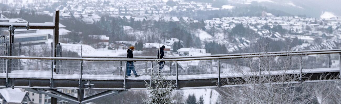 Spaziergänger gehen eine Brücke neben der Winterberger Bobbahn mit Blick auf die Landschaft im Schnee., © David Inderlied/dpa